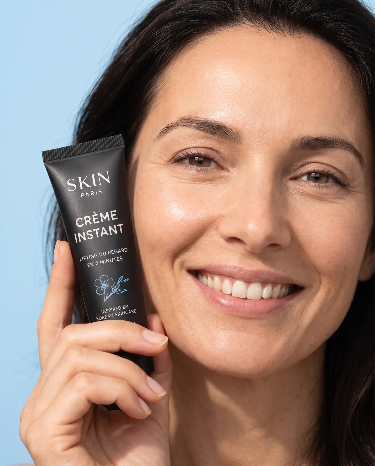 Une femme souriante au regard reposé tient le tube Crème Instant contre sa joue